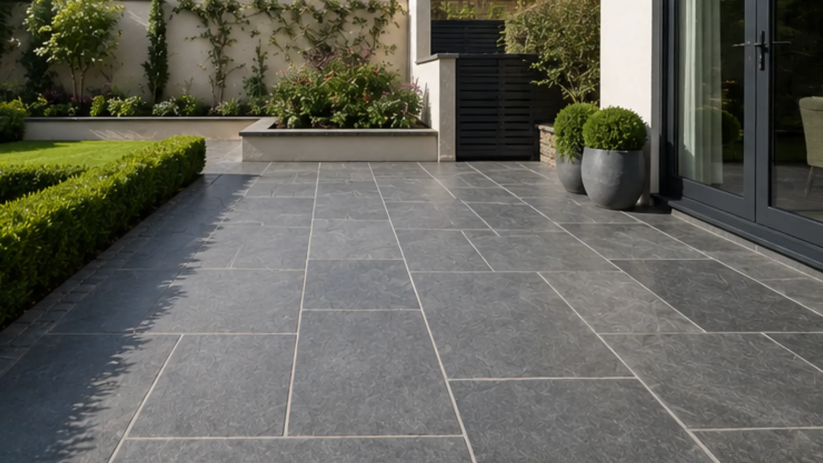 Dark natural stone patio beside bi-fold doors with trimmed hedging and raised planting bed